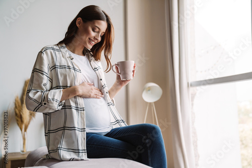 Fotografie Beautiful happy pregnant woman smiling and drinking coffee while sitting on couc