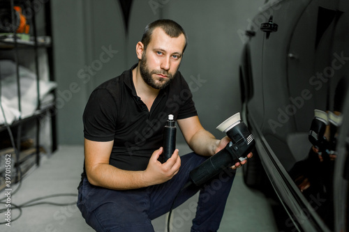 Car detailing series, polishing of luxury black car at auto service station. Handsome young bearded man worker, posing to camera with bottle of wax or cream and orbital polishing machine