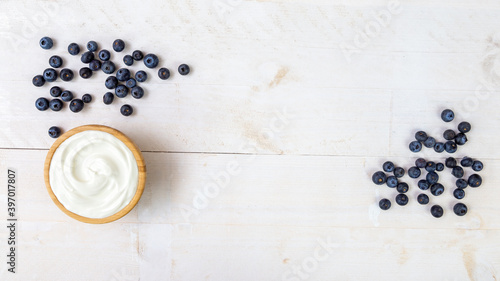 Heap of juicy blueberries and wooden bowl of smooth white yoghurt on white wooden kitchen table from directly above with copy space in the middle. Healthy breakfast ready to eat.