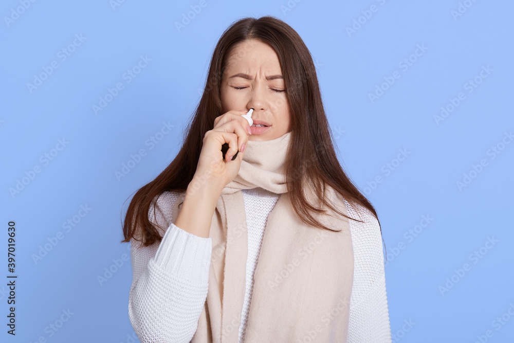 Young beautiful female with long brown hair keeping eyes closed and using nasal spray, wearing casual clothing and being wrapped in scarf, being infected with covid-19, standing against blue wall.