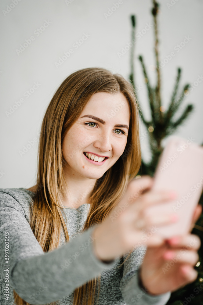 Happy woman talking on video call with relatives on the phone. Young girl are photographed or take selfie near the Christmas tree. Merry Christmas and Happy Holidays. Close up. Happy woman talking on video call with relatives on the phone. Young girl are photographed or take selfie near the Christmas tree. Merry Christmas and Happy Holidays. Close up.