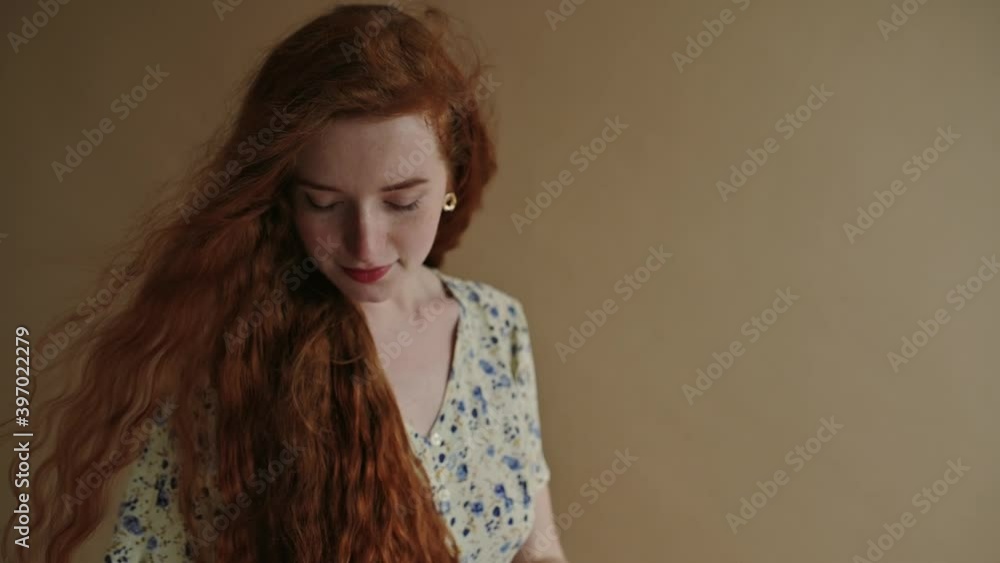 Hair Care conception: happy smiling redhead woman with long natural curly hair touching it. Model wearing floral print summer dress, posing on beige background. Slow Motion 100 fps Hair Care conception: happy smiling redhead woman with long natural curly hair touching it. Model wearing floral print summer dress, posing on beige background. Slow Motion 100 fps
