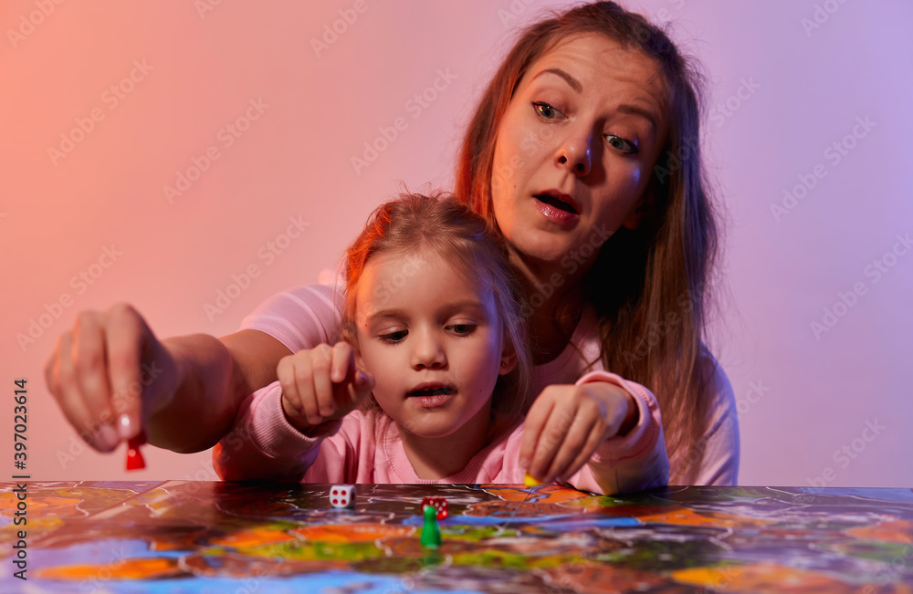 Fototapeta premium The family is having fun playing together at home in a board game. Board game field, many figures, coins and sandglass. Mother and cute little daughter play, holding cards joyful emotions on the face.
