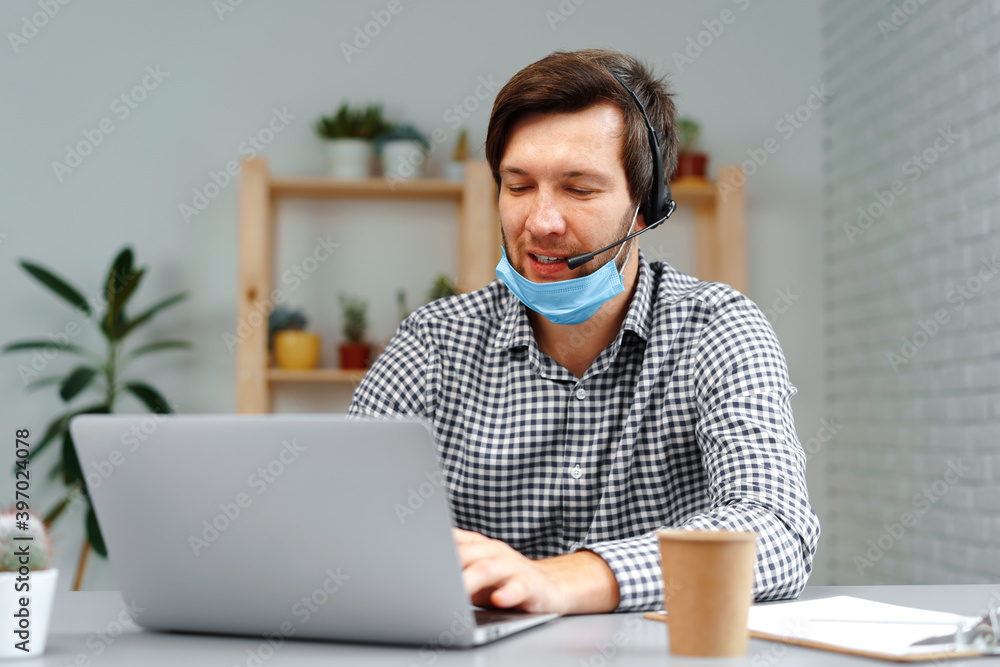 Young man working on laptop with headset and face mask at home