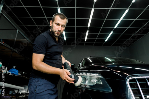 Car detailing series, polishing concept. Professional male auto service worker, wearing black t-shirt and pants, waxing and polishing headlight of black car with polish machine, looking at camera