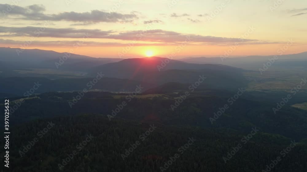 Lens flare in mountains at orange sunset in summer morning aerial view drone. Sun rays. Green pine forest slopes of mountain range. Bright disk of sun in blue sky slowly sets tops of mountains. Nature Lens flare in mountains at orange sunset in summer morning aerial view drone. Sun rays. Green pine forest slopes of mountain range. Bright disk of sun in blue sky slowly sets tops of mountains. Nature