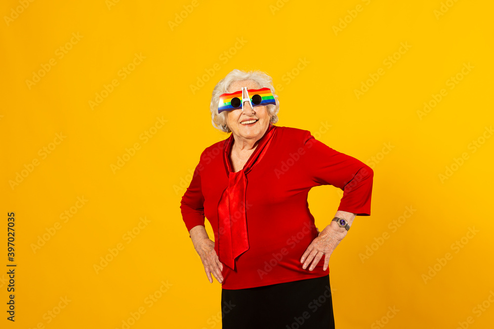 Studio portrait of a senior woman wearing a red shirt and rainbow sunglasses against a yellow background Studio portrait of a senior woman wearing a red shirt and rainbow sunglasses against a yellow background