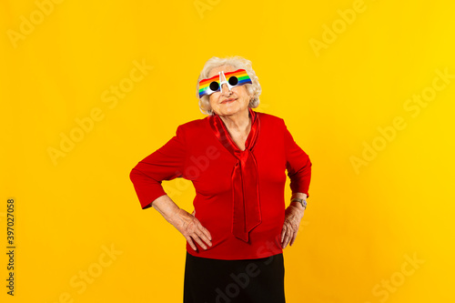 Studio portrait of a senior woman wearing a red shirt and rainbow sunglasses against a yellow background