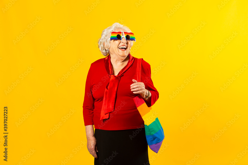 Studio portrait of a senior woman wearing a red shirt, rainbow sunglasses and a bag, against a yellow background Studio portrait of a senior woman wearing a red shirt, rainbow sunglasses and a bag, against a yellow background