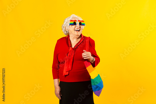 Studio portrait of a senior woman wearing a red shirt, rainbow sunglasses and a bag, against a yellow background © Jordi Salas