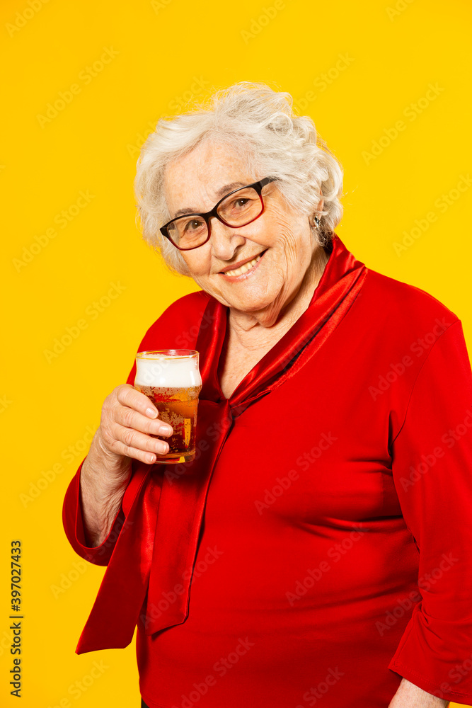 Studio portrait of a senior woman wearing red shirt with a glass of a half pint beer, against a yellow background Studio portrait of a senior woman wearing red shirt with a glass of a half pint beer, against a yellow background