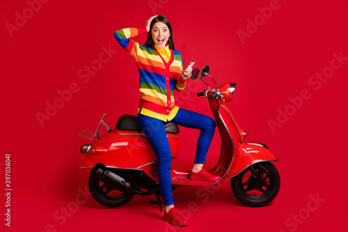 Photo portrait of shocked girl touching head holding phone in one hand sitting on scooter isolated on vivid red colored background