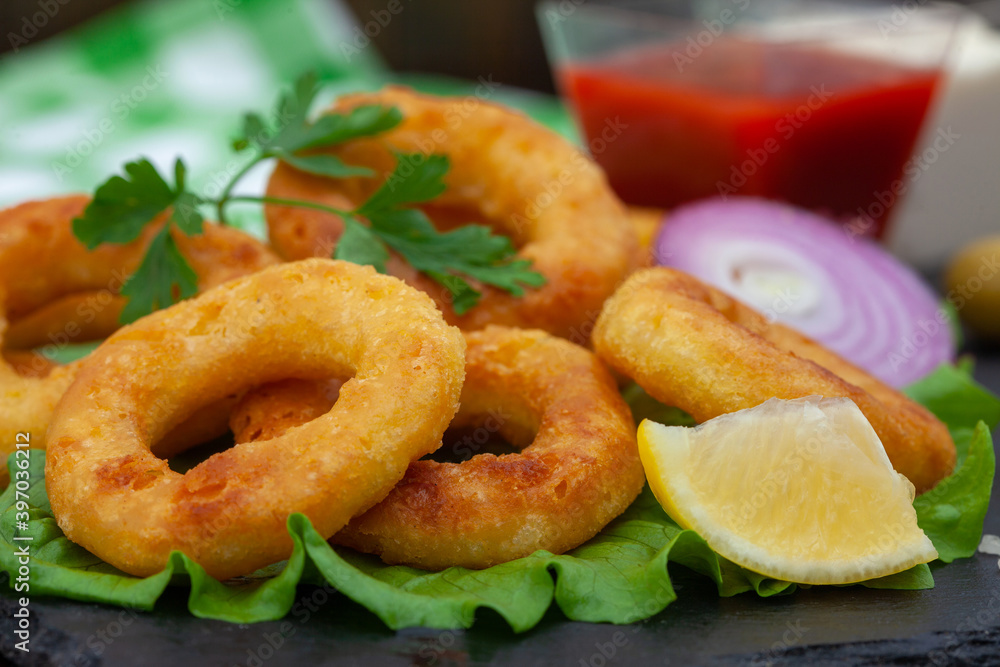 Deep fried squid or fish rings appetizer. Breaded squid or fish rings with lemon, ketchup and tartar sauce. Close up of golden, crispy calamari snack. Deep fried squid or fish rings appetizer. Breaded squid or fish rings with lemon, ketchup and tartar sauce. Close up of golden, crispy calamari snack.