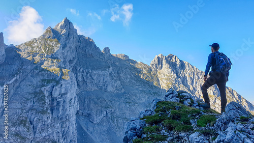 A man with a backpack standing in the shadow at the mountain peak and enjoying the view on higher  very steep peaks in front of him. Way to the Grimming in Austrian Alps. Freedom and exploration