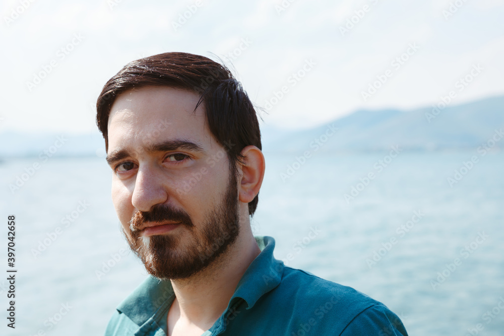 Portrait of calm handsome man with beard and peaceful sight standing against sea and mountains and looking at camera Portrait of calm handsome man with beard and peaceful sight standing against sea and mountains and looking at camera
