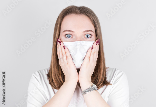 Young woman doctor in a medical mask looks at the camera with surprise,holding her face with her hands. Сoncept of covid 19, flu and seasonal cold.