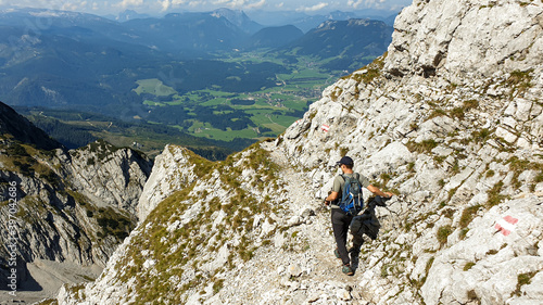 A man walking on a very narrow and steep pathway along a mountain wall in Austrian Alp, leading to the top of Grimming. There is a small city in the bottom of the valley. Dangerous trail. Adventure