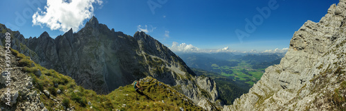 A woman sitting at the edge of a mountain with a view on a vast valley. There are sharp mountains and high peaks around. The Alpine slopes are almost barren. Lush green valley. Bright day. Freedom.