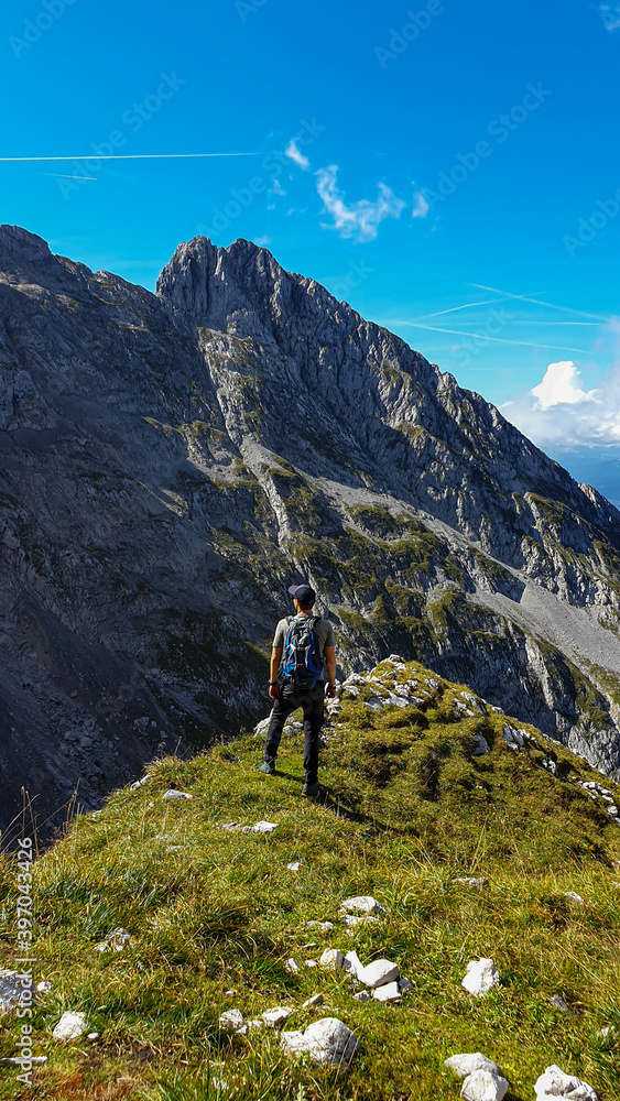 A man standing at the edge of a mountain with a view on high mountain behind her. There are sharp mountains and high peaks around. The Alpine meadow is overgrown with lush green grass. Adventure