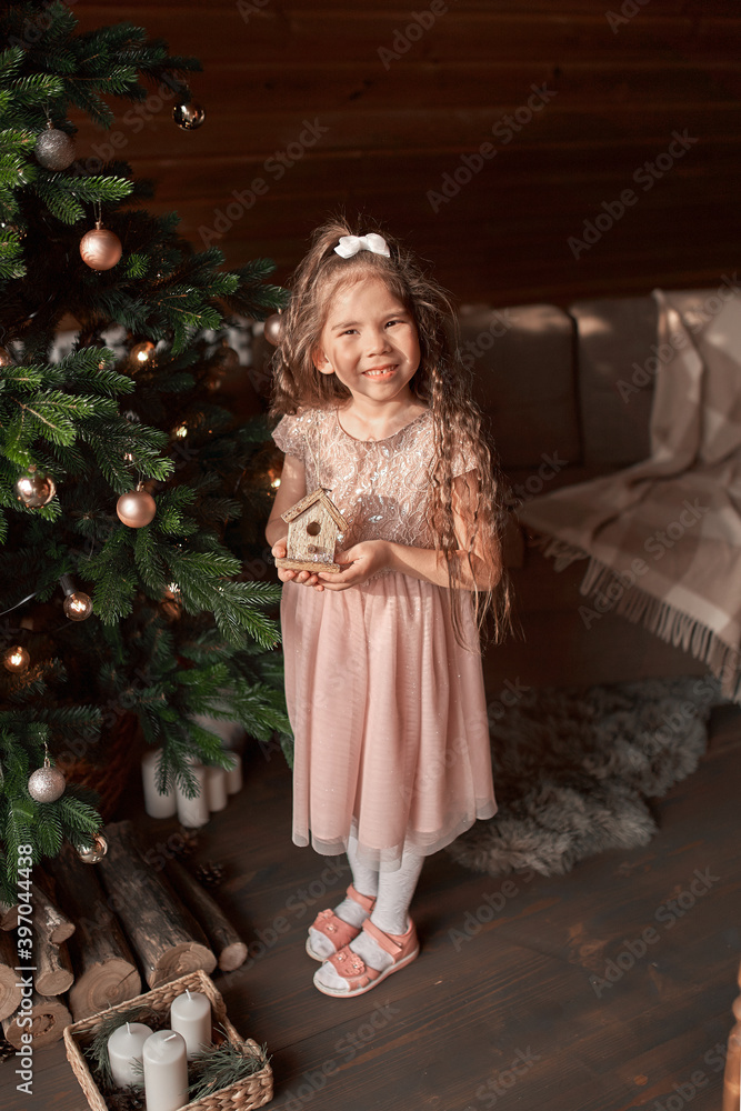 Adorable little toddler girl with curly hair in a warm knitted dress decorating a beautiful Christmas tree at a big window with a view of a snowy garden