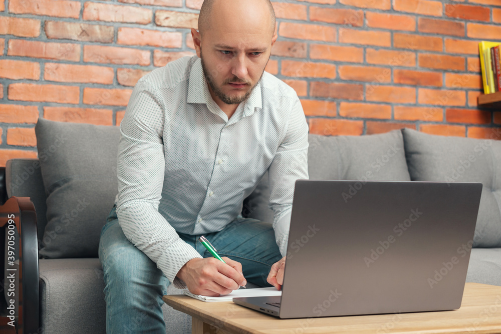Bearded bald attractive businessman or freelancer sitting on a sofa and working on laptop from home, modern interior loft design Bearded bald attractive businessman or freelancer sitting on a sofa and working on laptop from home, modern interior loft design