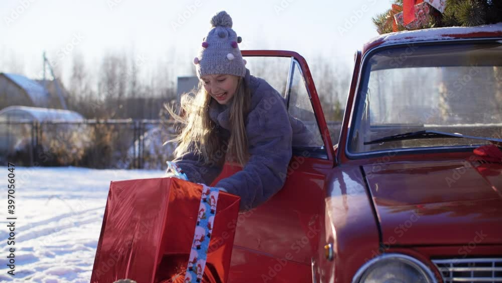 Joyful teen girl holding big gift box with red ribbon inside car on Christmas eve. Happy girl taking New Year present sitting in red car on holiday morning. Happy winter holiday concept Joyful teen girl holding big gift box with red ribbon inside car on Christmas eve. Happy girl taking New Year present sitting in red car on holiday morning. Happy winter holiday concept