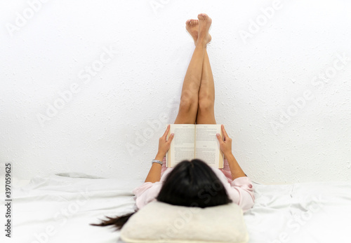 Young woman with legs up on the wall lying on bed and reading book