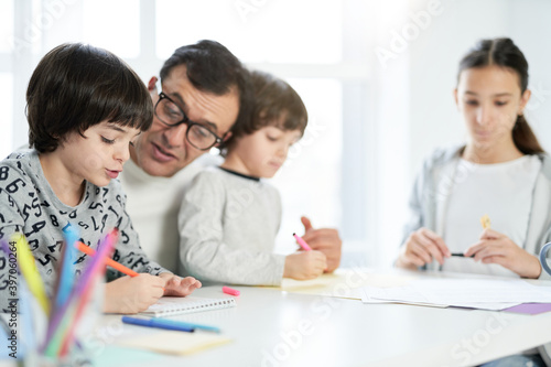 Learning process. Adorable little latin boy drawing something while spending time with his father and siblings at home. Businessman working from home and watching children