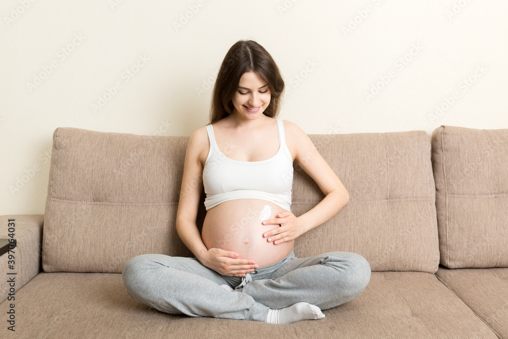 a pregnant girl sits at home on the bed and smears an anti-stretch mark cream on her stomach. Pregnancy, motherhood, preparation and expectation concept a pregnant girl sits at home on the bed and smears an anti-stretch mark cream on her stomach. Pregnancy, motherhood, preparation and expectation concept