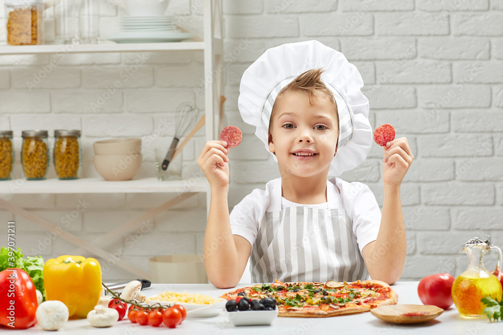 little boy in chef hat and an apron cooking pizza in the kitchen. the child holding sausage slices. having fun little boy in chef hat and an apron cooking pizza in the kitchen. the child holding sausage slices. having fun