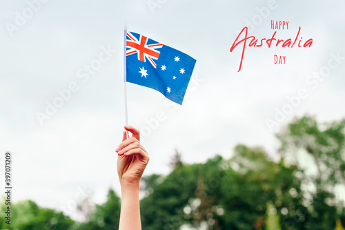 Happy Australia Day card with greeting text. Closeup of woman human hand arm waving Australian flag against blue sky. Proud citizen man celebrating national Australia Day in January outdoors. © anoushkatoronto