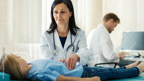 Female doctor in white coat palpating belly of girl lying on couch and talking to little patient, male doctor making notes in medical history at desk in background