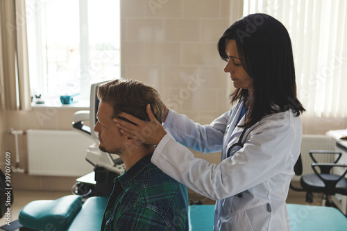 Middle aged female doctor in white coat turning slightly head of male patient to check his injured neck