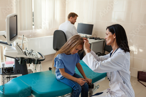 Female pediatrician in white coat checking blonde hair of little girl sitting on couch in doctors office  male doctor working at desk in background
