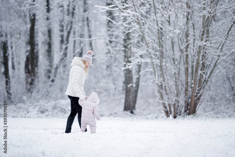 Naklejka premium Little daughter in overalls and young adult mother walking on white first snow at park. Spending time together in beautiful winter day. Enjoying peaceful stroll.