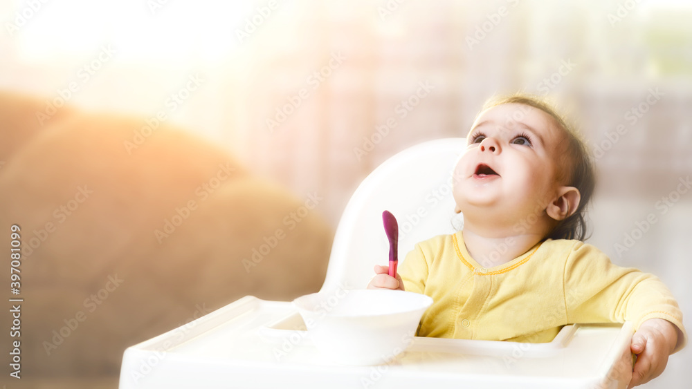 Little baby sitting in high chair and eating food. baby's first feeding. 6 month old todler holding spoon and plate and looking up Little baby sitting in high chair and eating food. baby's first feeding. 6 month old todler holding spoon and plate and looking up