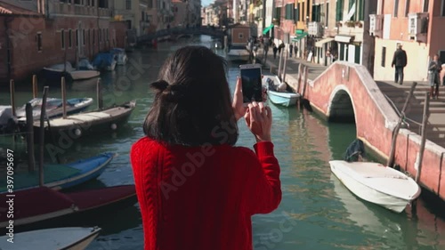 Rear view of beautiful asian girl in red sweater taking photo of the picturesque venetian canal with her smartphone from edge of the bridge. Charming woman spending her vacation in Venice.