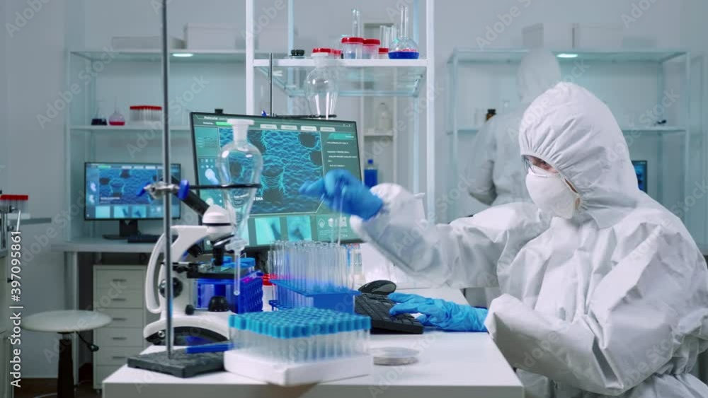 Biochemists in coverall typing at pc evolution of vaccine composition in equipped laboratory analysing test tube with sample. Doctors examining virus evolution using high tech researching diagnosis Biochemists in coverall typing at pc evolution of vaccine composition in equipped laboratory analysing test tube with sample. Doctors examining virus evolution using high tech researching diagnosis