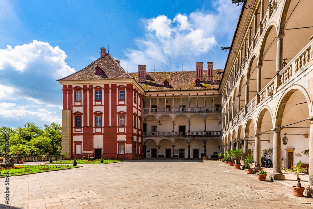 Opocno, Czech Republic - June 16 2020: View of the castle courtyard with arcades and red facade. Green lawn with statue and flowers in foreground. Sunny summer day with blue sky and white clouds.