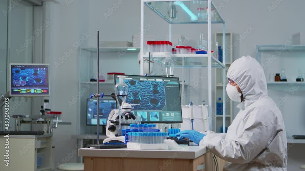 Chemist in ppe suit typing on computer checking virus development in equipped laboratory. Team doctors working with various bacteria, tissue and blood samples, pharmaceutical research for antibiotics Chemist in ppe suit typing on computer checking virus development in equipped laboratory. Team doctors working with various bacteria, tissue and blood samples, pharmaceutical research for antibiotics
