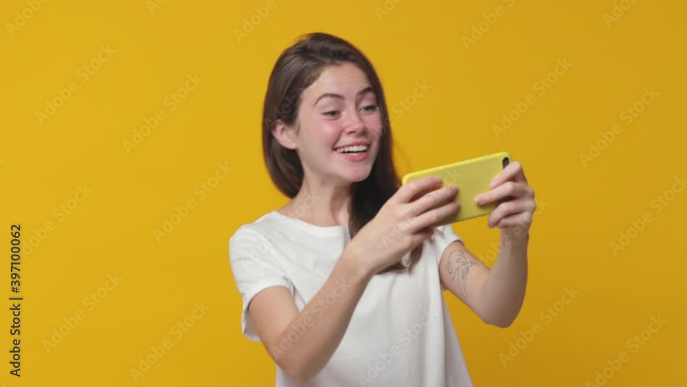 Excited cheerful upset brunette young woman 20s years old wearing casual white t-shirt posing isolated on yellow color background in studio. People lifestyle concept. Playing game on mobile cell phone Excited cheerful upset brunette young woman 20s years old wearing casual white t-shirt posing isolated on yellow color background in studio. People lifestyle concept. Playing game on mobile cell phone