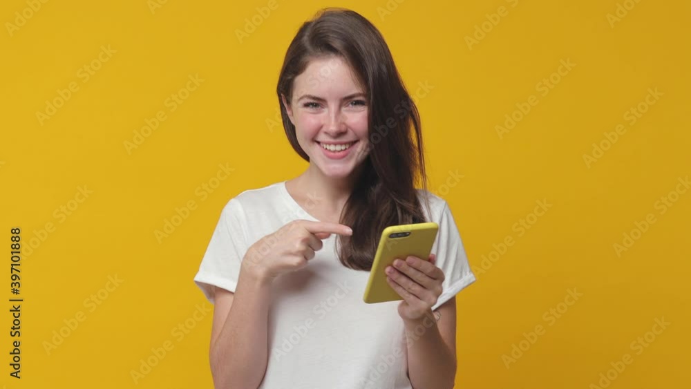 Surprised brunette young woman 20s years old in white t-shirt isolated on yellow background in studio. People lifestyle concept. Pointing index finger on mobile cell phone typing sms message say wow Surprised brunette young woman 20s years old in white t-shirt isolated on yellow background in studio. People lifestyle concept. Pointing index finger on mobile cell phone typing sms message say wow