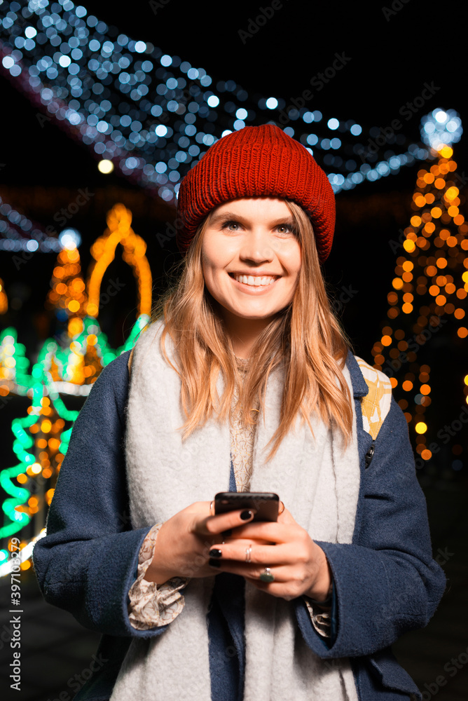 Young pretty lady is smiling at the camera while holding her phone outdoor at night with Christmas lights.