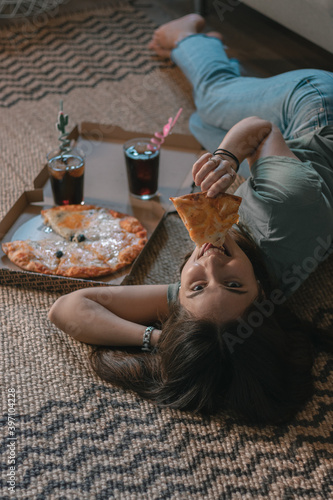 Pretty girl eating pizza from the box and drinking drinks lying on the carpet on the floor of the house, happy, with loose hair, barefoot and casual clothes.