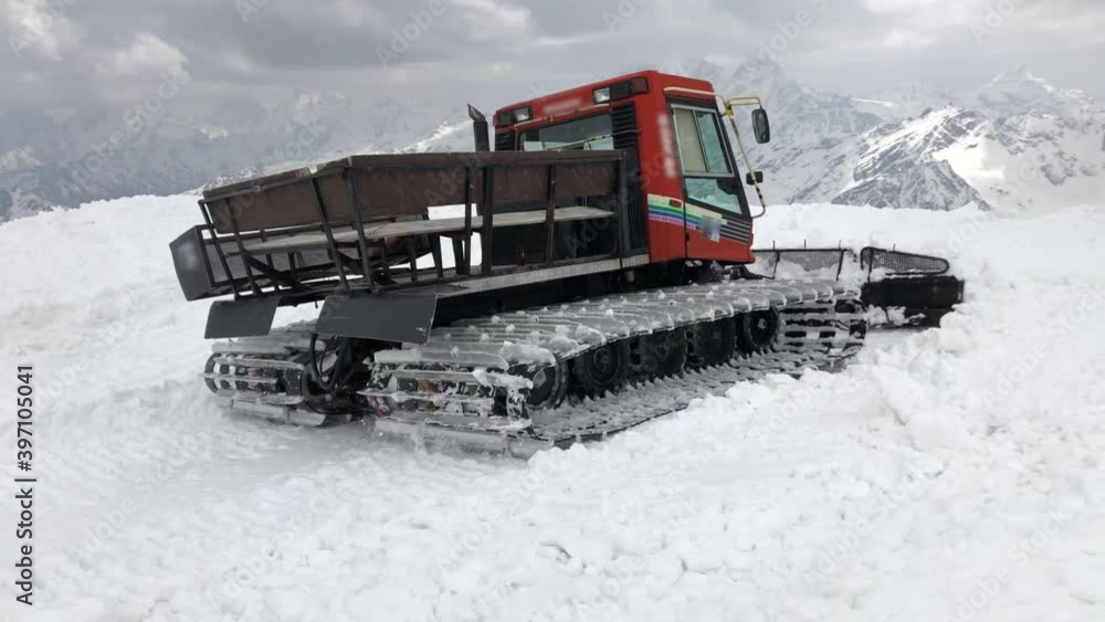 Close-up work of wide snowcat rides on snow and rakes it against the backdrop of snow-capped mountains. concept of preparing snow slopes for the ski season and snow harvesting Close-up work of wide snowcat rides on snow and rakes it against the backdrop of snow-capped mountains. concept of preparing snow slopes for the ski season and snow harvesting