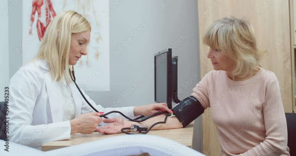 Side view. Portrait of Caucasian beautiful female doctor in white coat sitting in clinic cabinet measuring blood pressure of senior woman patient with medical manometer at appointment. Health concept Side view. Portrait of Caucasian beautiful female doctor in white coat sitting in clinic cabinet measuring blood pressure of senior woman patient with medical manometer at appointment. Health concept