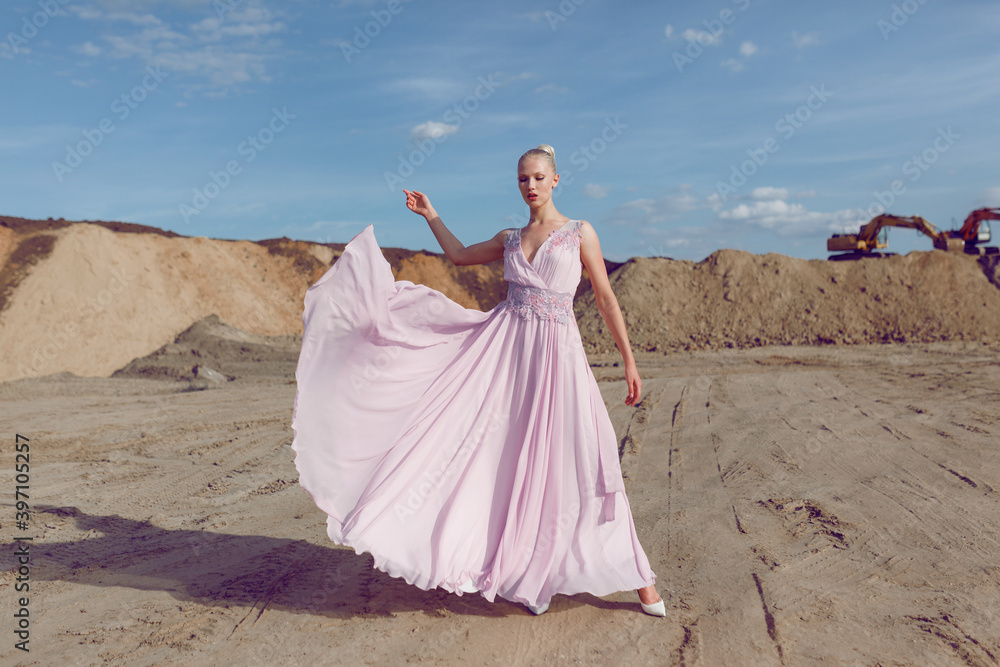 Obraz premium Full length portrait of a blonde high girl in fashionable flying pink maxi dress poses in sand quarry on sunny day.