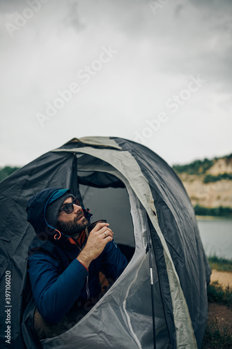 Young handsome bearded man sitting in a tent, drinking coffee and looking at sky.
