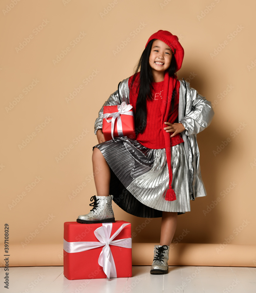 Asian brunette is leaning her foot on a large gift box, holding a gift in one hand and pointing to an empty space for text with the other. Gift concept for Christmas, New Year or birthday.