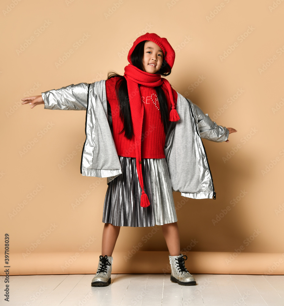 Asian little model girl in a red beret, scarf, sweater and silver skirt and jacket posing with pleasure on a beige background in the studio. Style concept for the modern child.